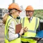 Construction workers wearing safety vests and helmets discussing plans at construction site.